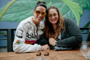 Two smiling women at a table, one in a colorful jacket and sunglasses, the other with glasses and a cardigan, with glass tumblers in front and a hop mural behind.