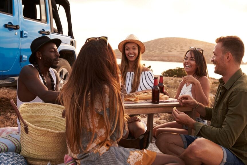 Group of friends enjoying food and drinks outdoors by a blue jeep during sunset.