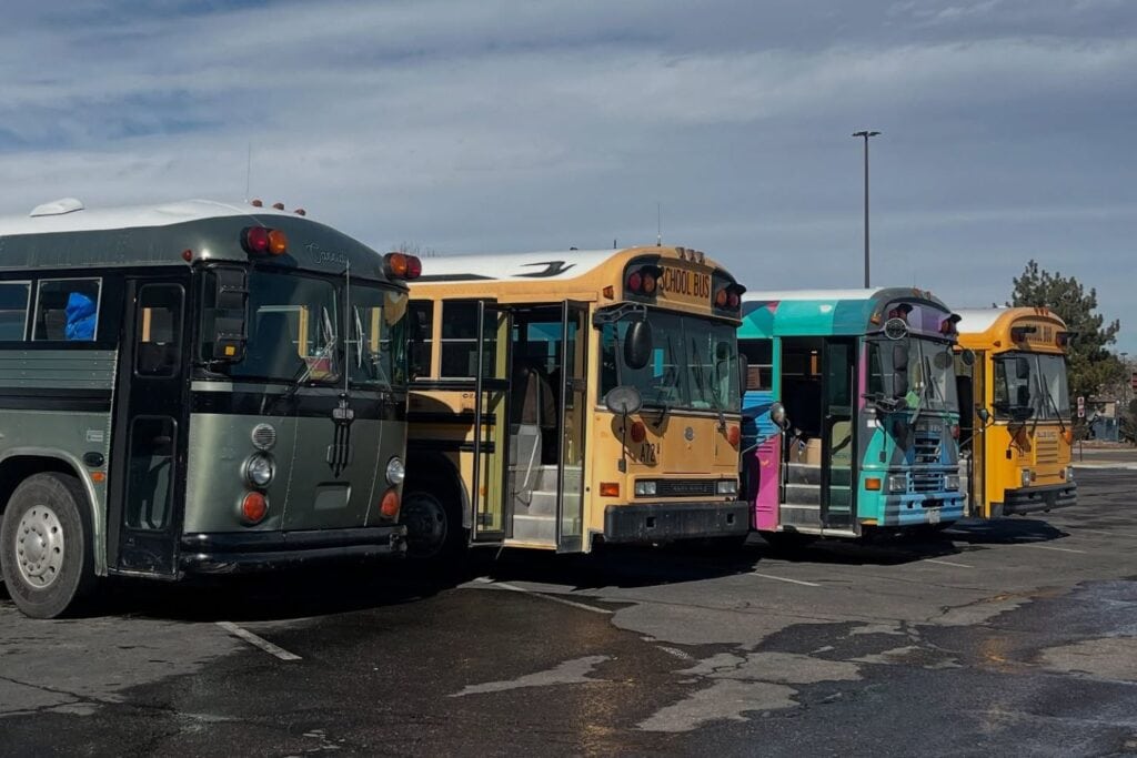 Row of colorful, modified school buses and coaches used for a "Bus to Show" service, parked in a lot under a partly cloudy sky.