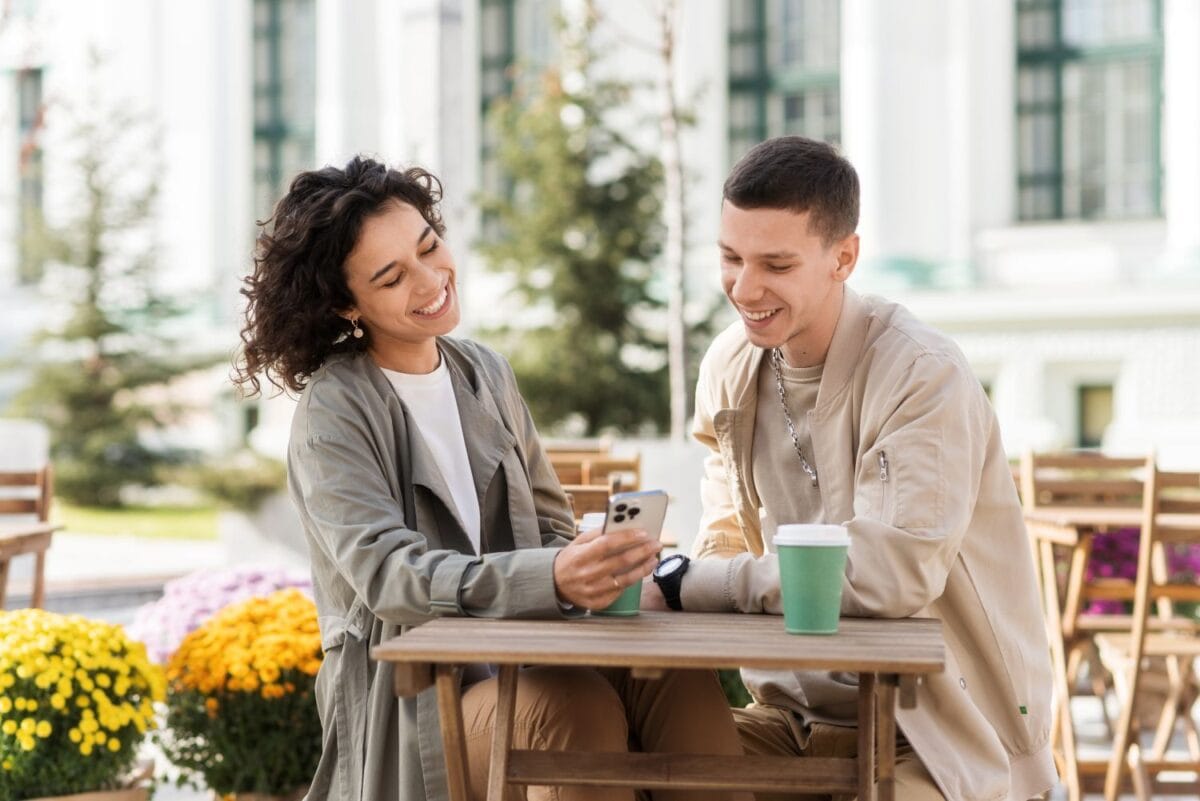 Top 12 Denver Date Ideas | A happy couple outdoors near a cafe. Looking in the phone, smiling, talking, coffee on the table. Autumn atmosphere